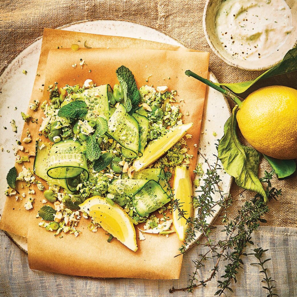 Fresh green coloured vegetable salad with shaved cucumber ribbons, herbs and chopped nuts on parchment paper, served with lemon wedges and creamy dressing.