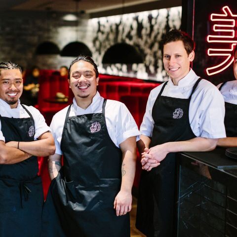 Four young men in aprons stand in a restaurant with Chinese characters in the background.