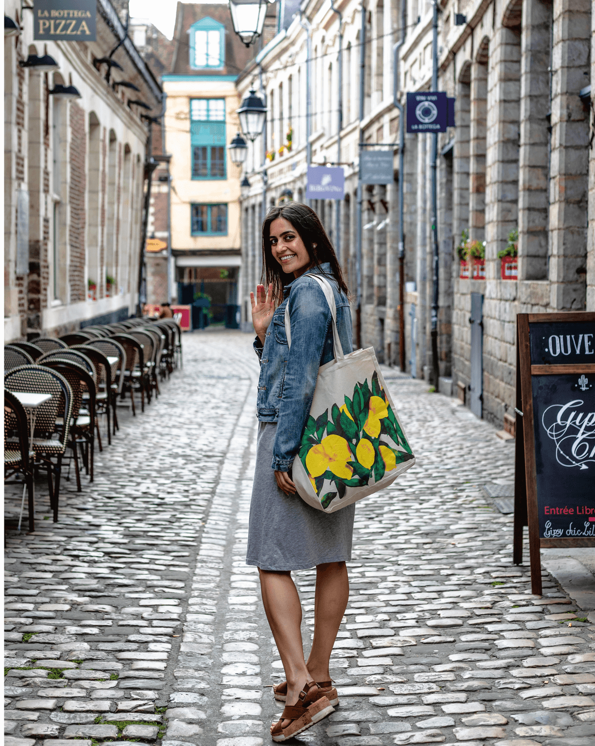 A young woman, Hannah Sunderani, waves to the camera on a cobblestone street.