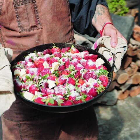 A man holding a cast-iron dish filled with roasted strawberries, ricotta and mint