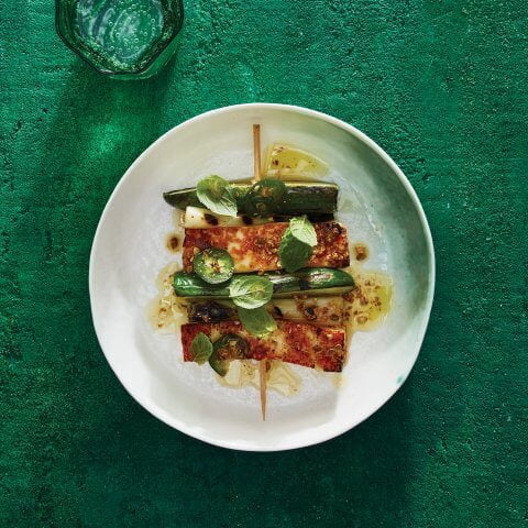 Skewered cucumbers and halloumi cheese on a white dish against a bright green table.