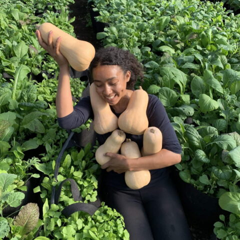 A woman kneels in a field and poses with an armful of squash.