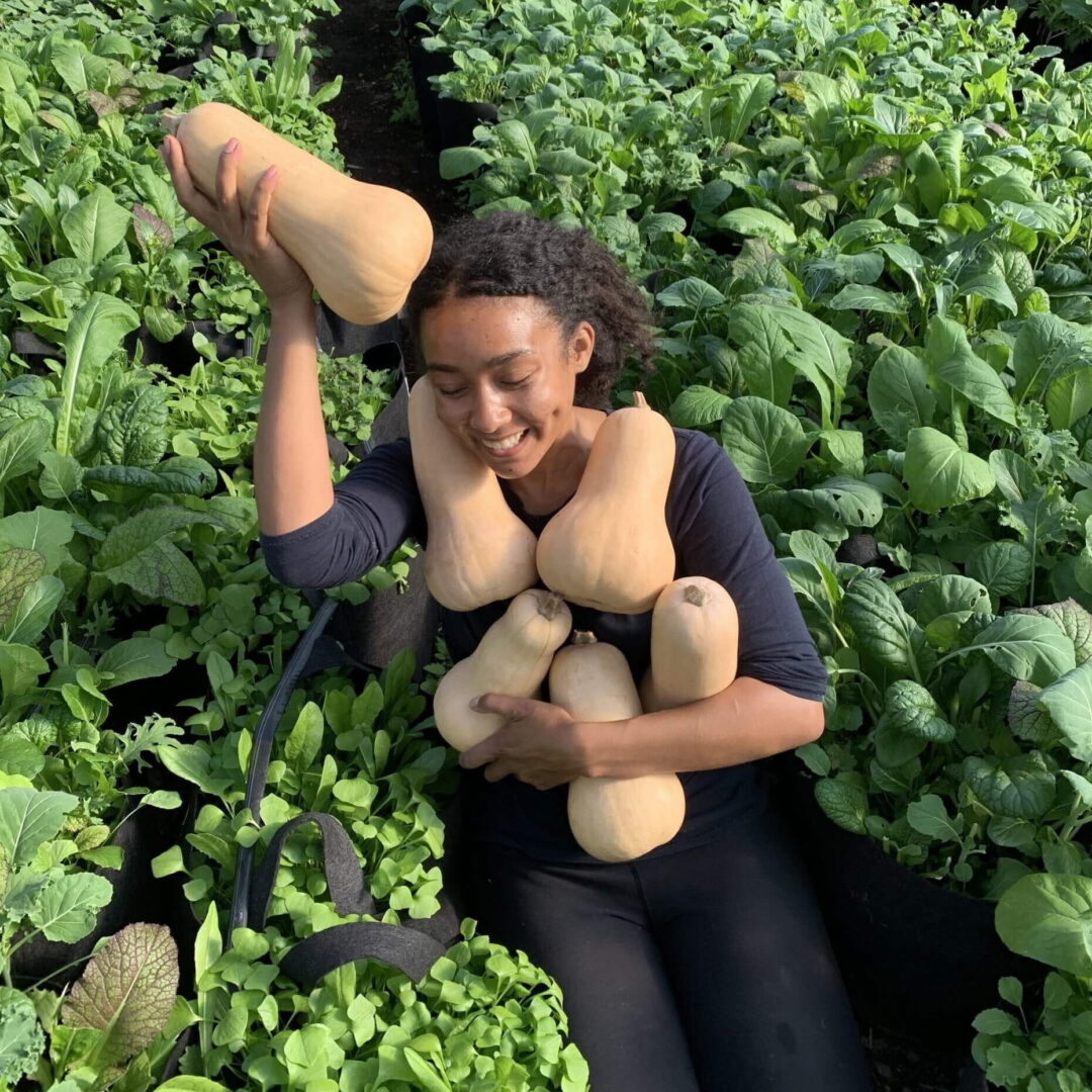 A woman kneels in a field and poses with an armful of squash.