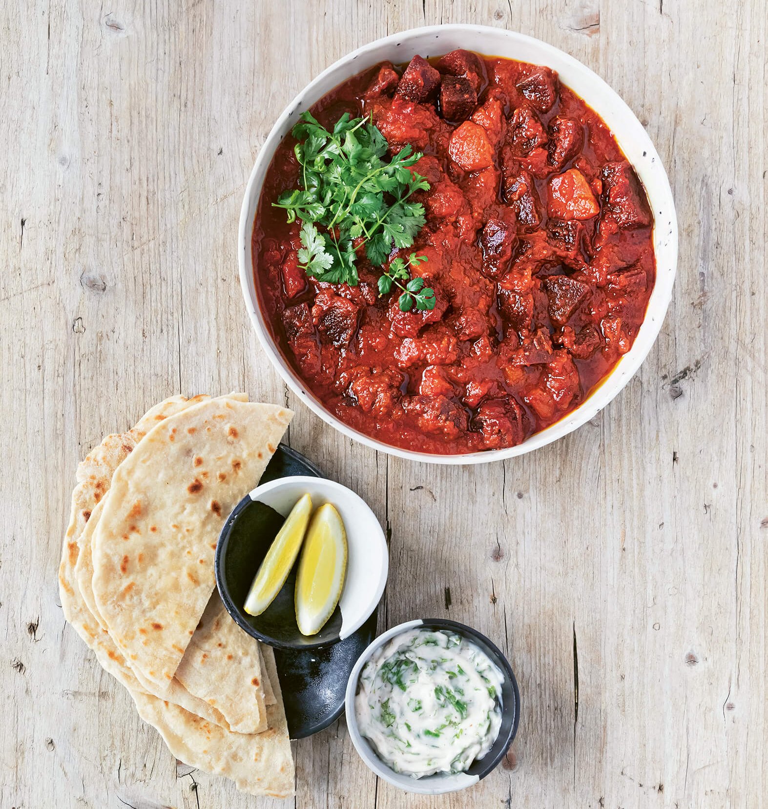 A bowl of curry with bread, lemons and dip on the side