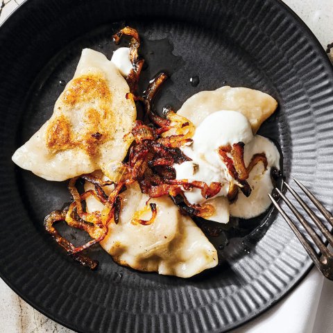 A black plate on a marble surface with pierogi, sour cream, bacon, onion and a fork