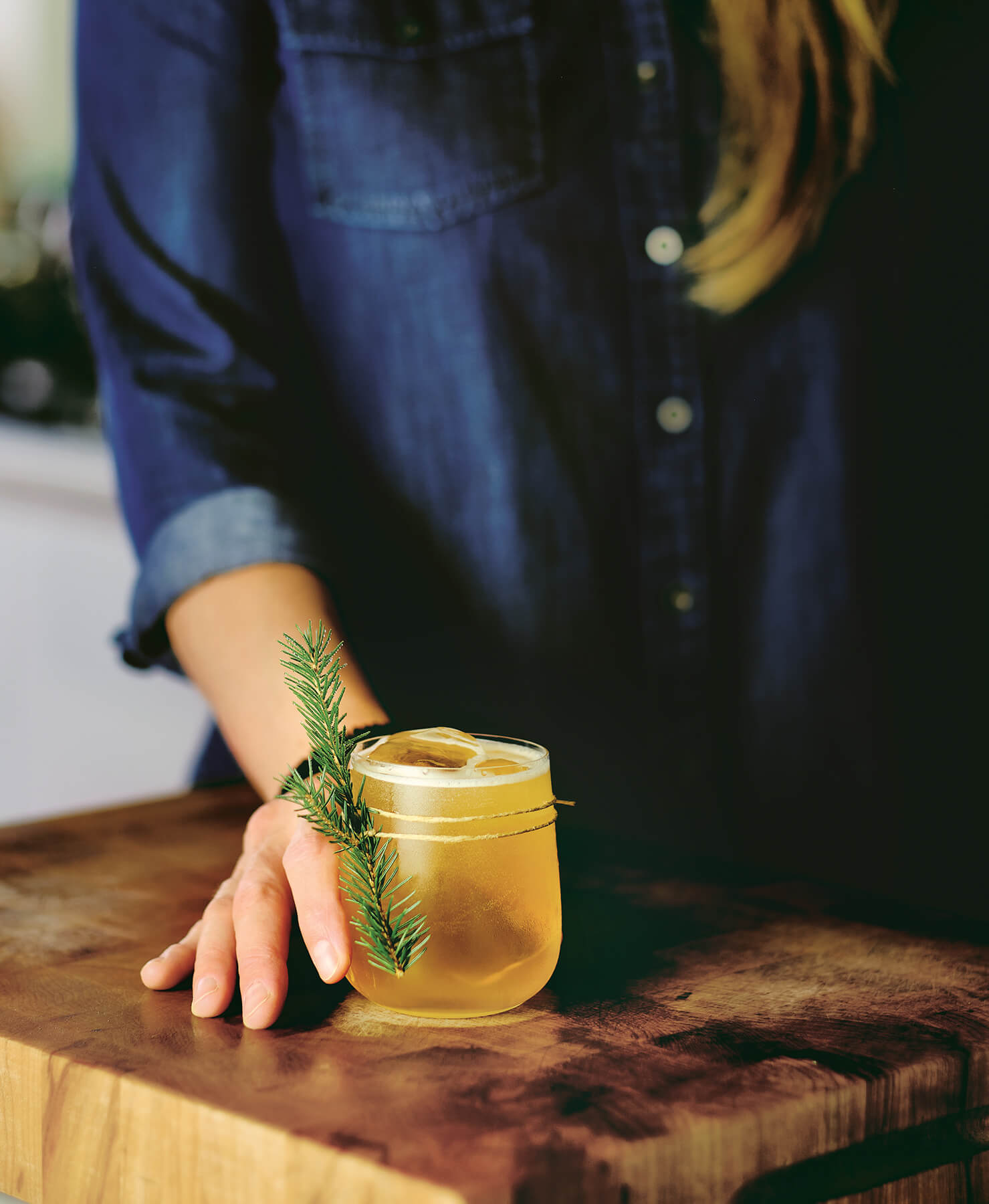 A woman holds an amber drink with an evergreen garnish