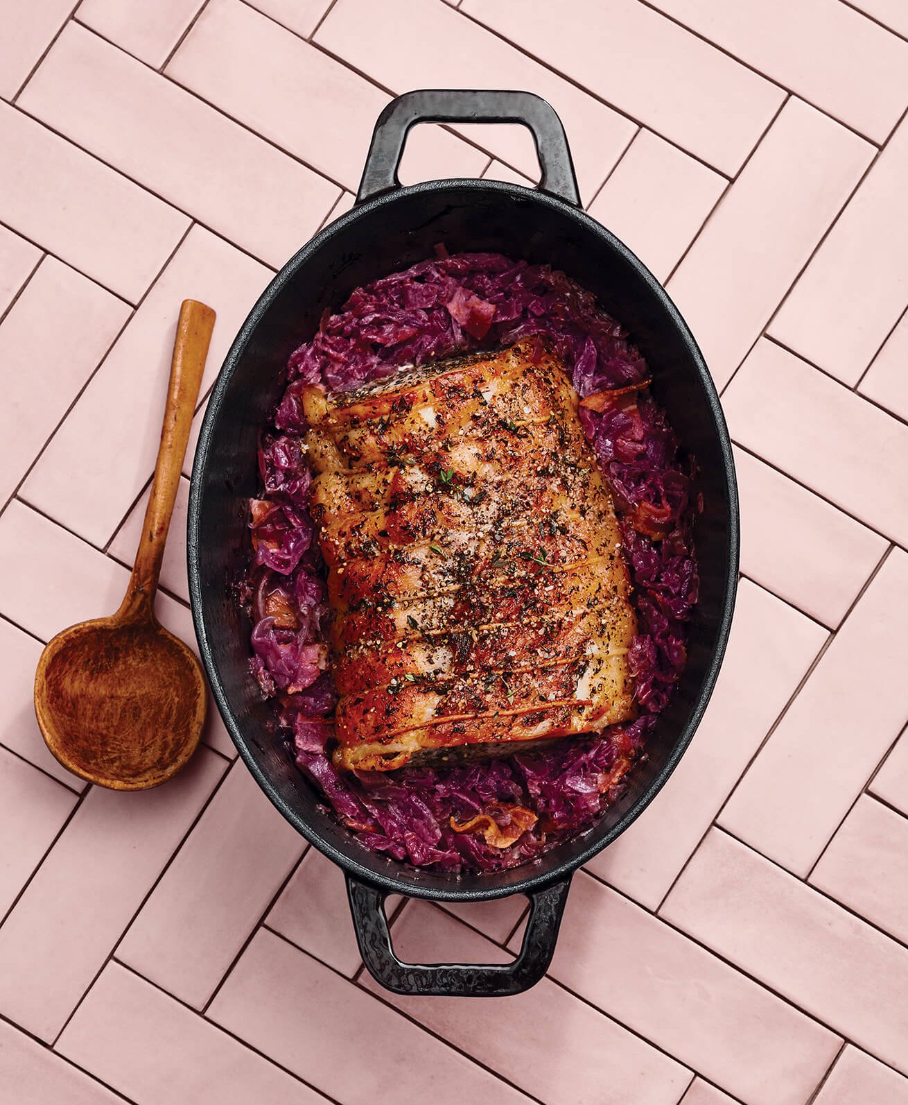 A cast iron dish with pork and cabbage and a wooden spoon on a pink countertop