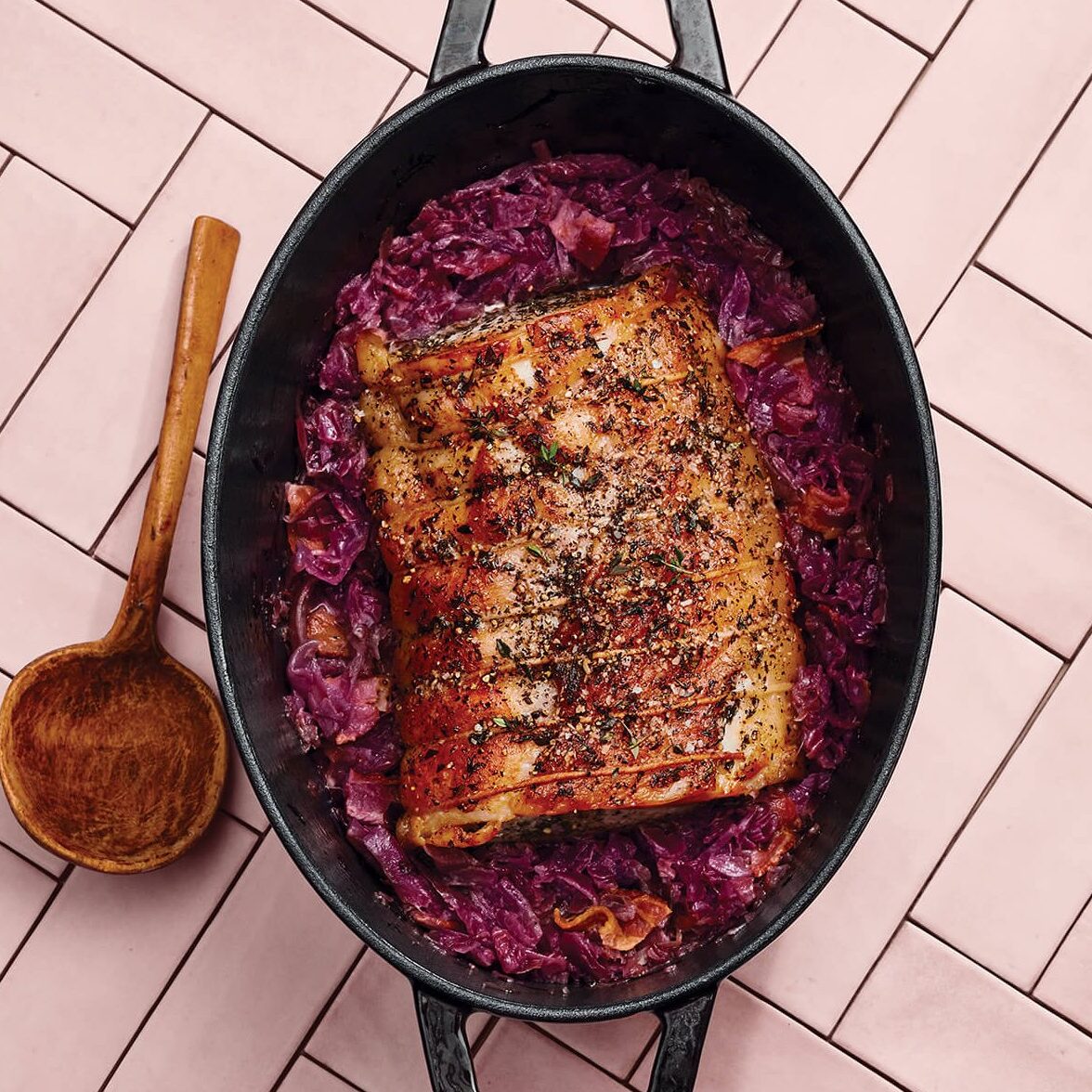A cast iron dish with roasted pork and cabbage with a wooden spoon on a pink countertop