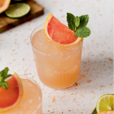 Glasses with a light pink drink and grapefruit garnish on a linoleum countertop.