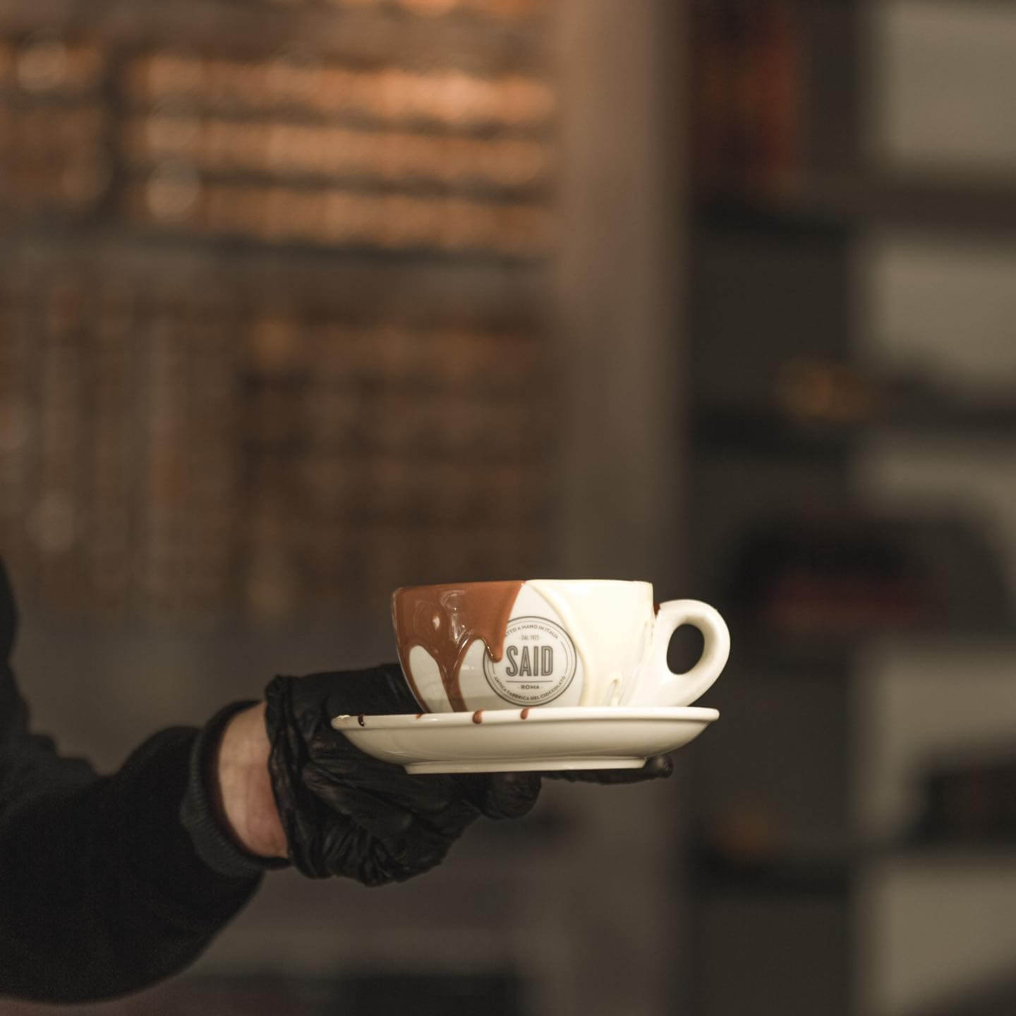 A gloved hand holds up a mug with chocolate overflowing.