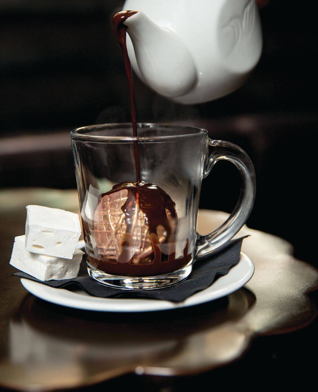 A white teapot pouring chocolate over a hot chocolate bomb with marshmallows on the side.