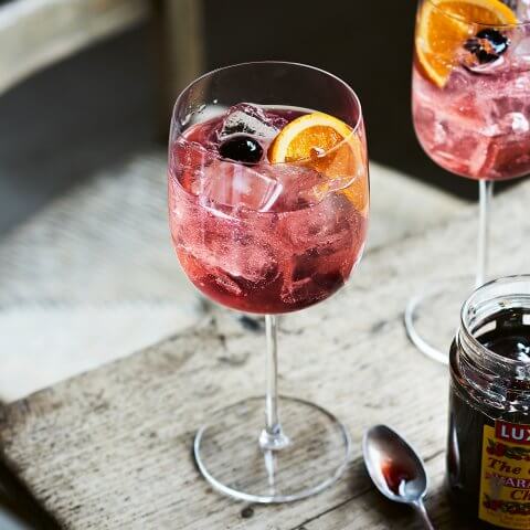 Two cocktails in wine glasses with orange wheel garnishes and a jar of maraschino cherries next to it on a grey wooden table