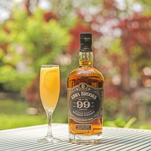 A bottle of Ezra Brooks bourbon and a champagne glass with an orange cocktail on a table outdoors with trees in the background.