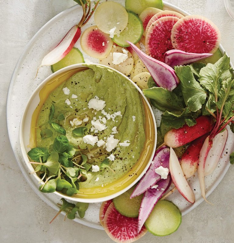 A white tabletop with a white plate laid with vegetables and a green dip.