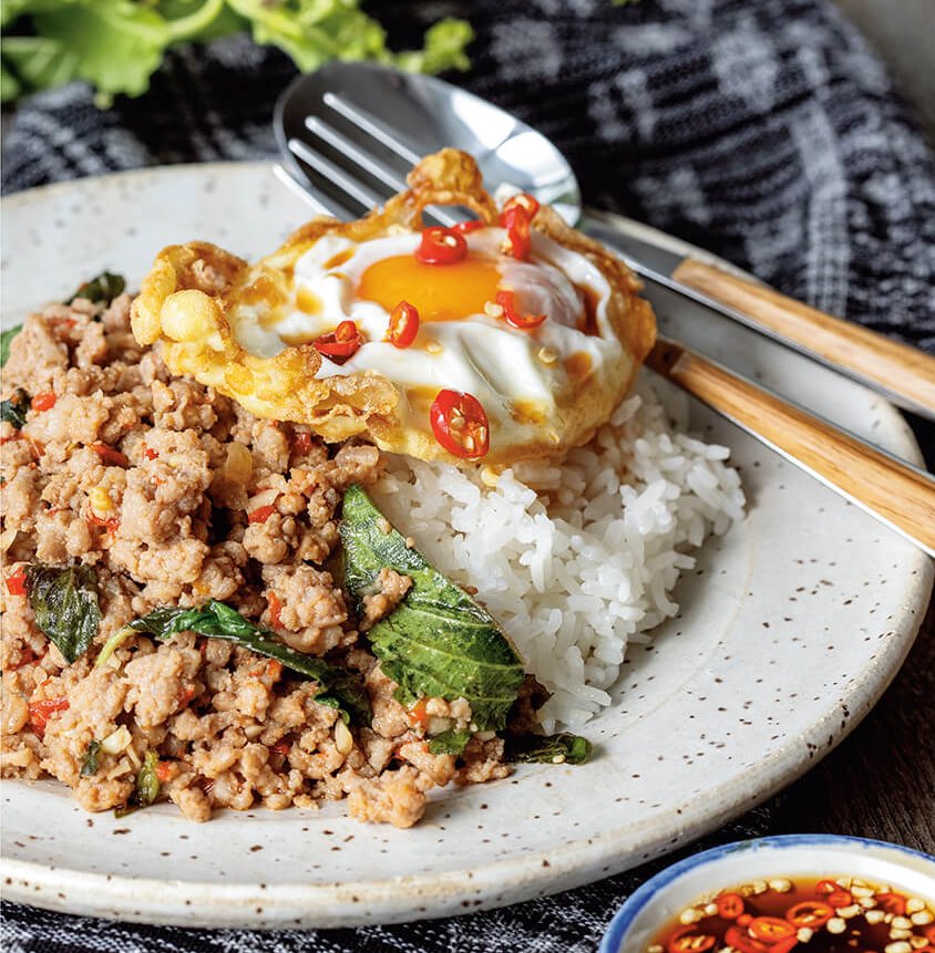 A dish with pad gaprao, a fork and spoon on a grey wooden tablecloth with blue linens and lettuce seen in the background.