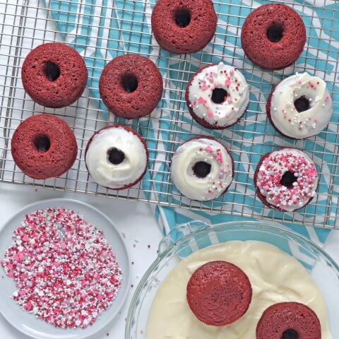 A cooling rack with red doughnuts in various stages of decoration on a blue cloth.