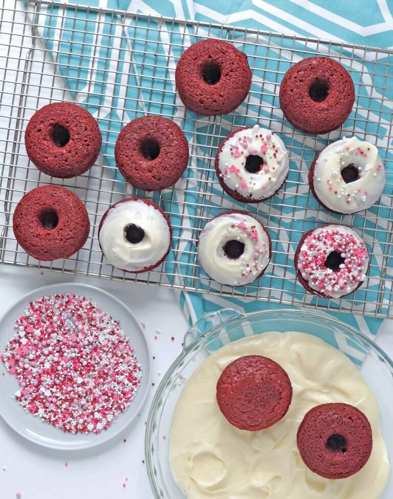 A cooling rack with red doughnuts in various stages of decoration on a blue cloth.