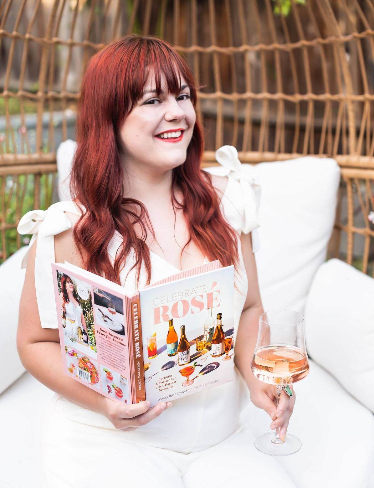 A woman sitting in a white outdoor chair holding a glass of rose wine and an open book