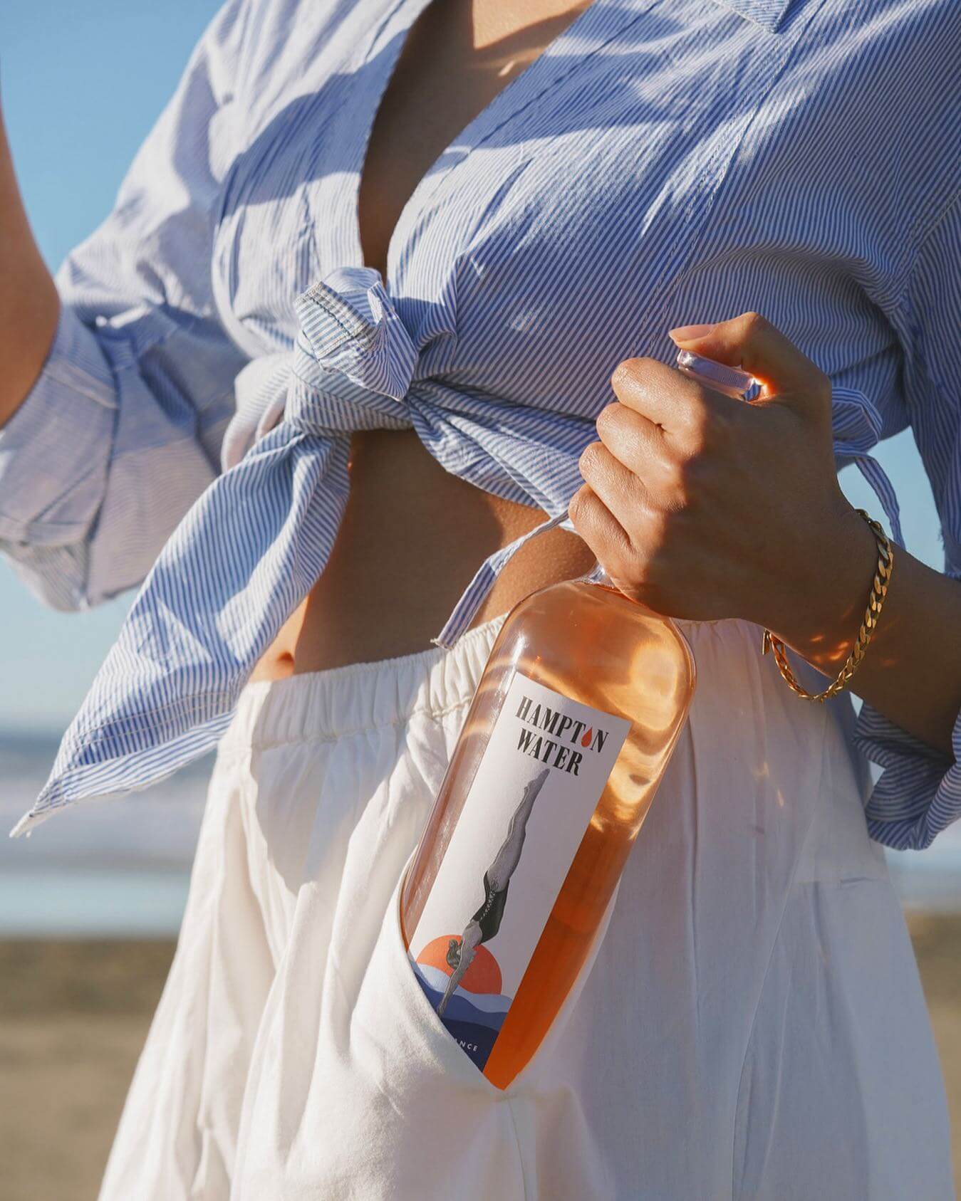 A woman in a white skirt and blue blouse holding a bottle of Hampton Water rose wine.