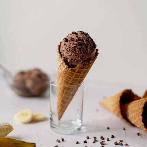 A chocolate ice cream cone resting in a glass with sliced bananas and other ingredients scattered nearby