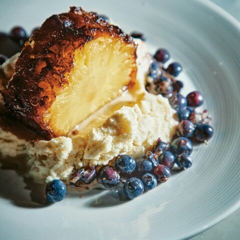 A slice of roasted pineapple surrounded by blueberries on a blue dish