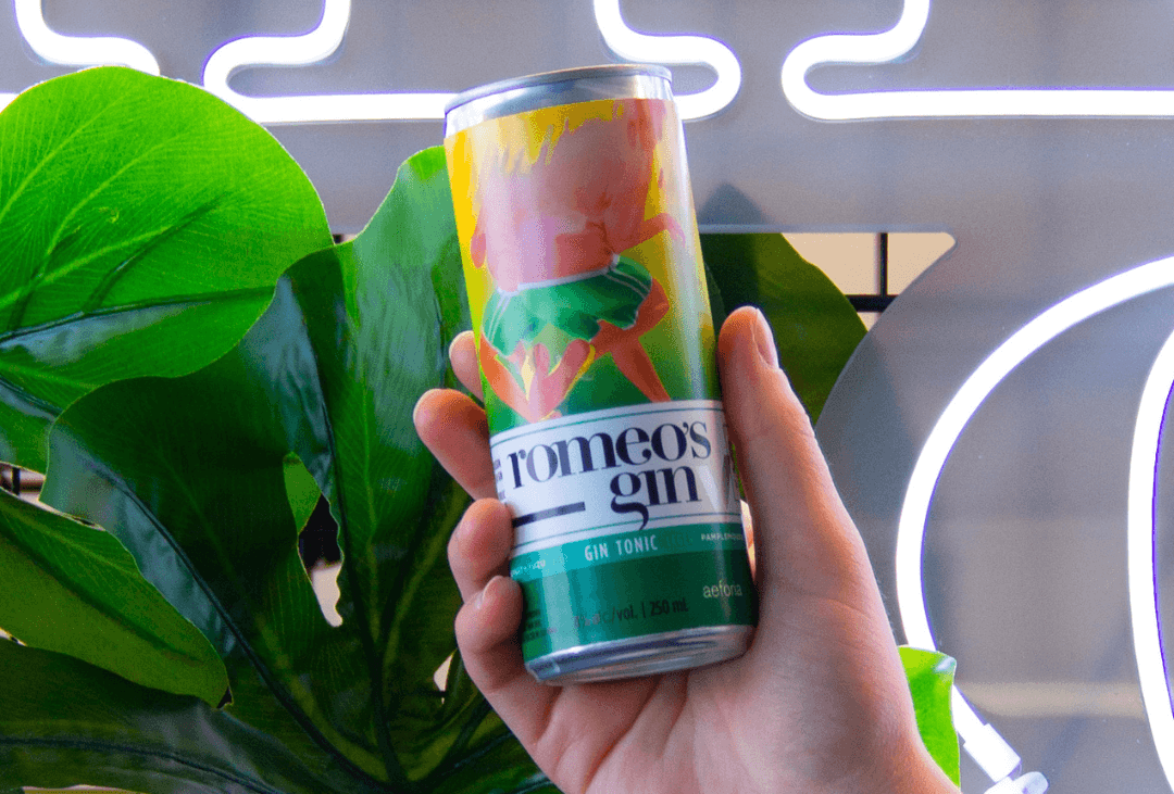 A person's hand holding a colourful drink can in front of green leaves and neon signage