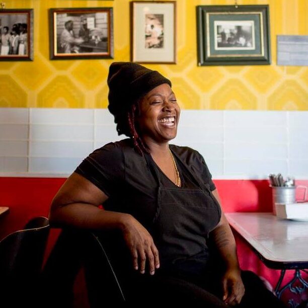 Jamaican-Canadian chef Suzanne Barr sitting at a table in a dining room with yellow walls covered in photos.