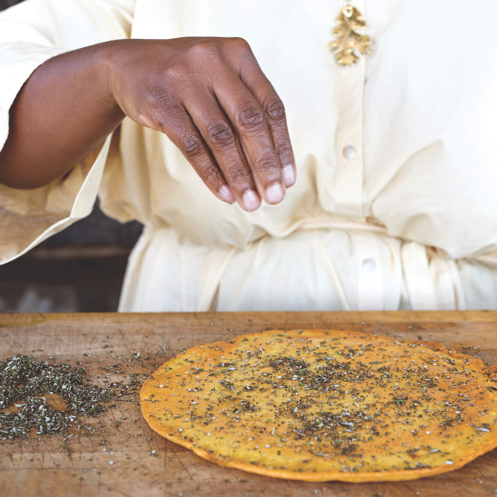 A woman smiles while she sprinkles a flat, round piece of bread with seasonings.