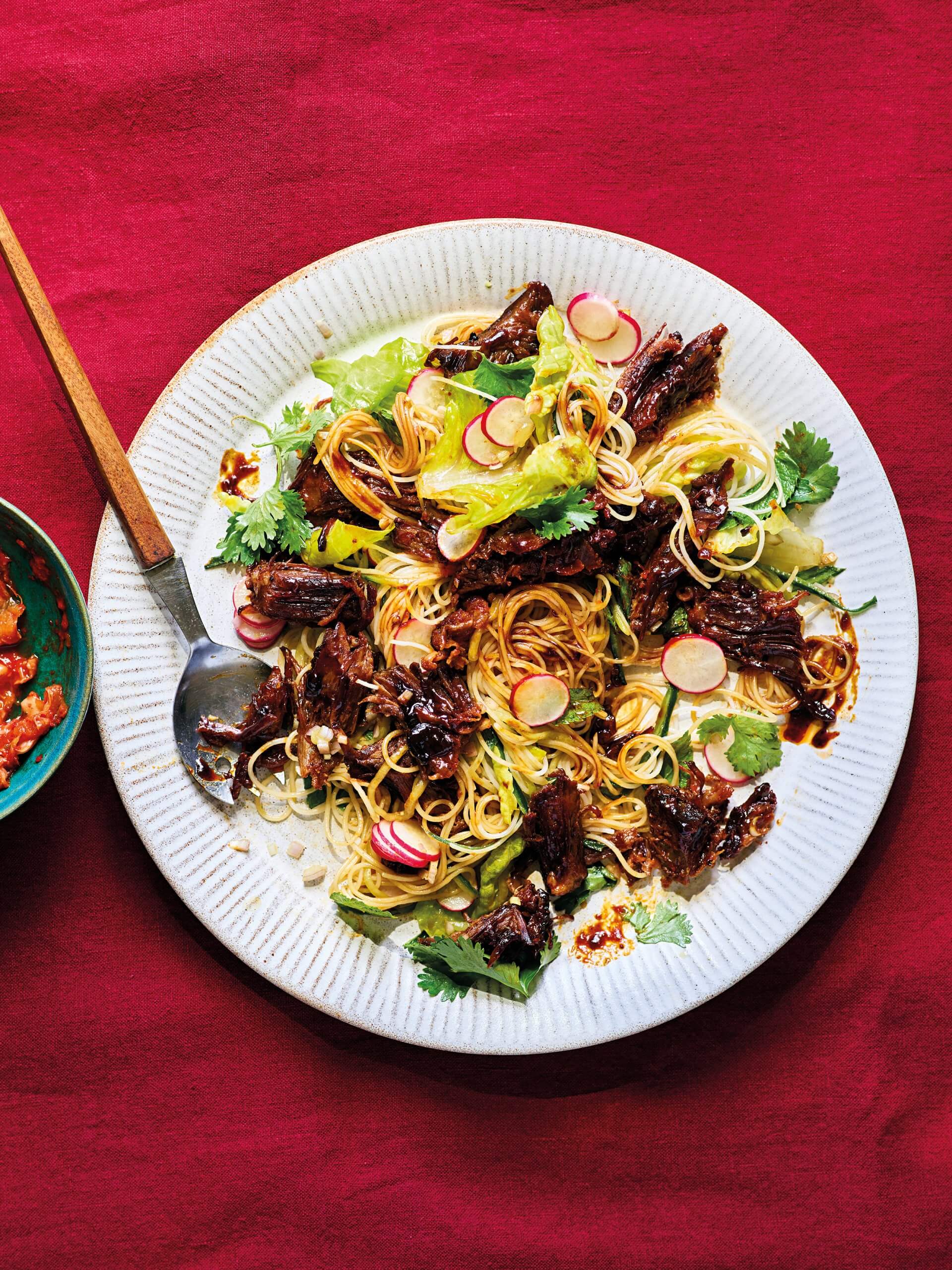 A white plate with rice noodle salad and short ribs on a bright red surface