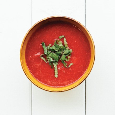 A small bowl of bright red soup with a lettuce garnish on bright white planks
