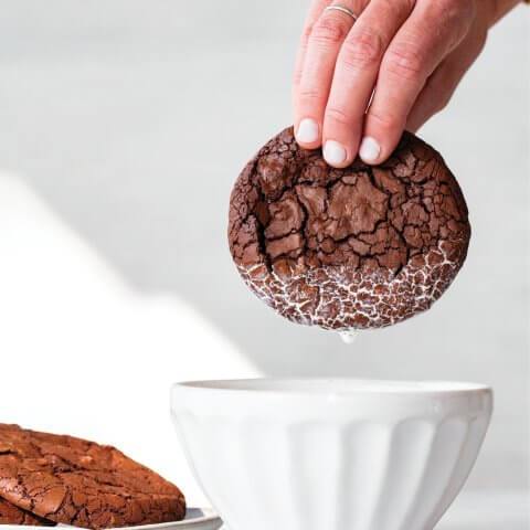 A person's hand dunking a chocolate cookie in a white bowl