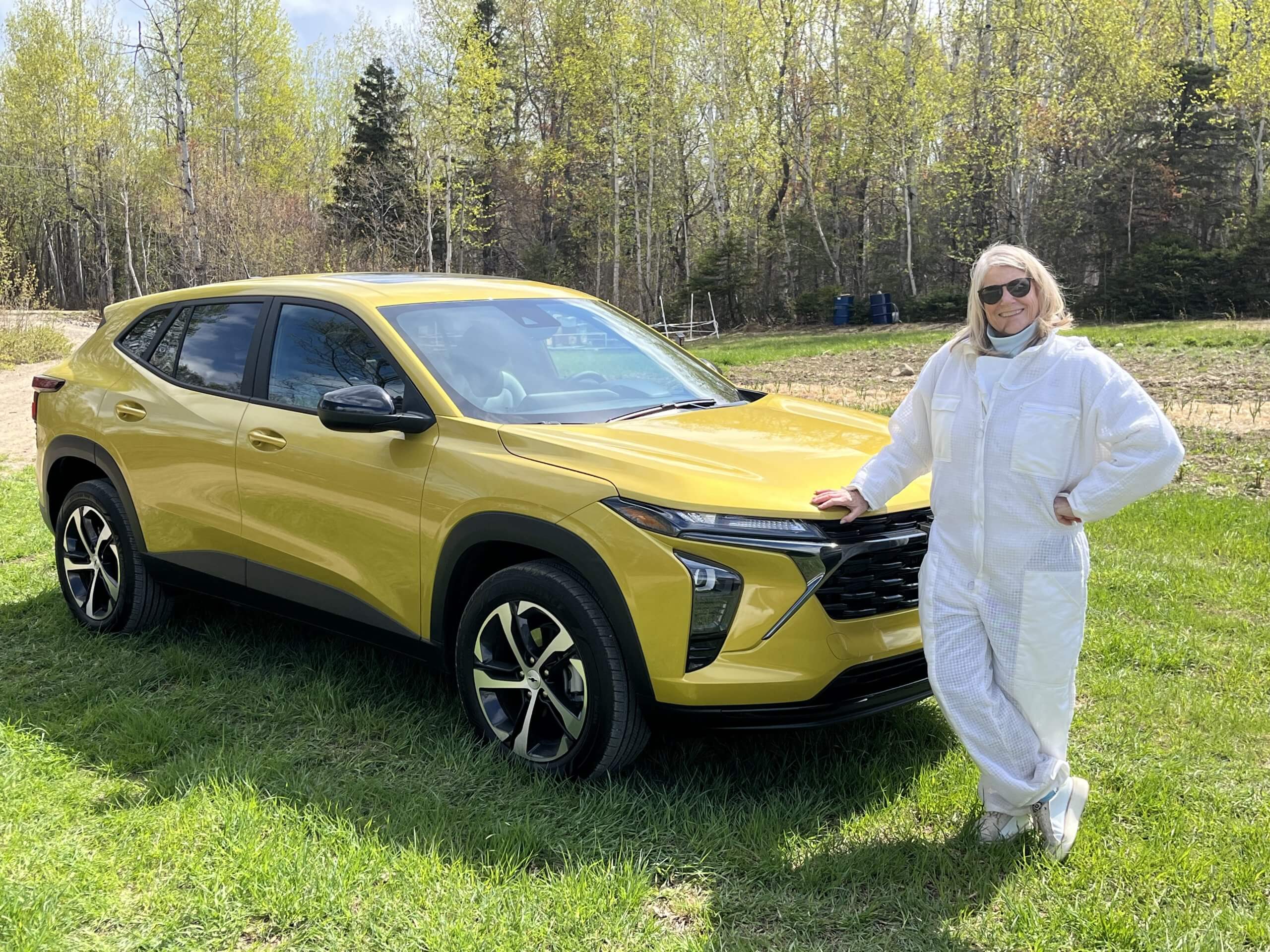 A woman, Jacqueline Howe, wears a white beekeeping suit and leans on a yellow SUV parked in a field