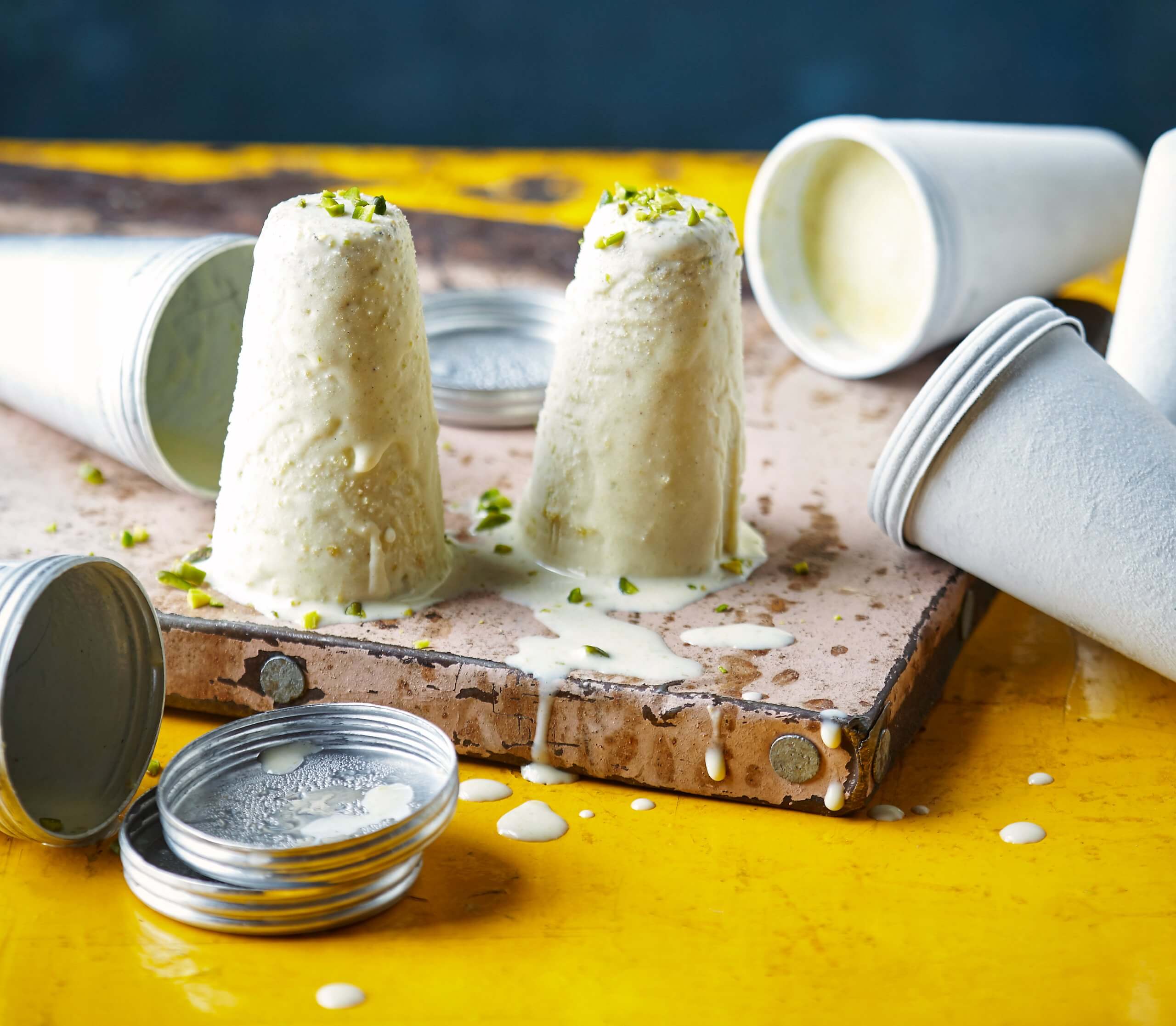 Two melting servings of Pistachio Kulfi paper molds in the background on a bright yellow surface