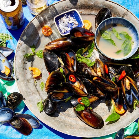 A plate of mussels on a bright blue tabletop, with salt and broth served on the side