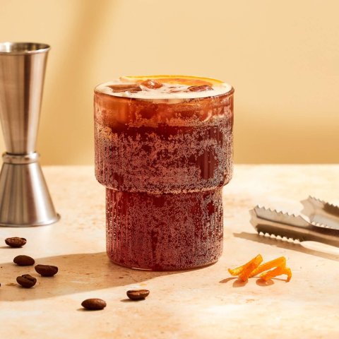A clear glass with a coffee cocktail, a silver bar measure, tongs, coffee beans and an orange peel rind on a yellow-ish background