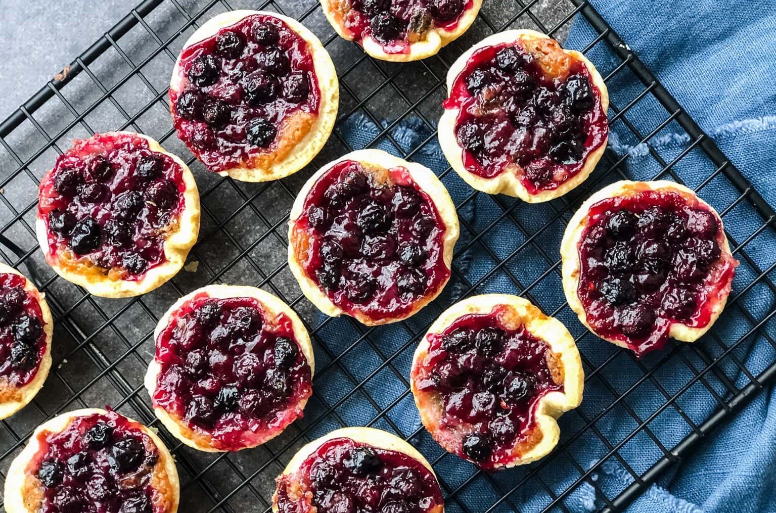 A rack of tarts topped with Saskatoon berries on a grey surface with a blue cloth