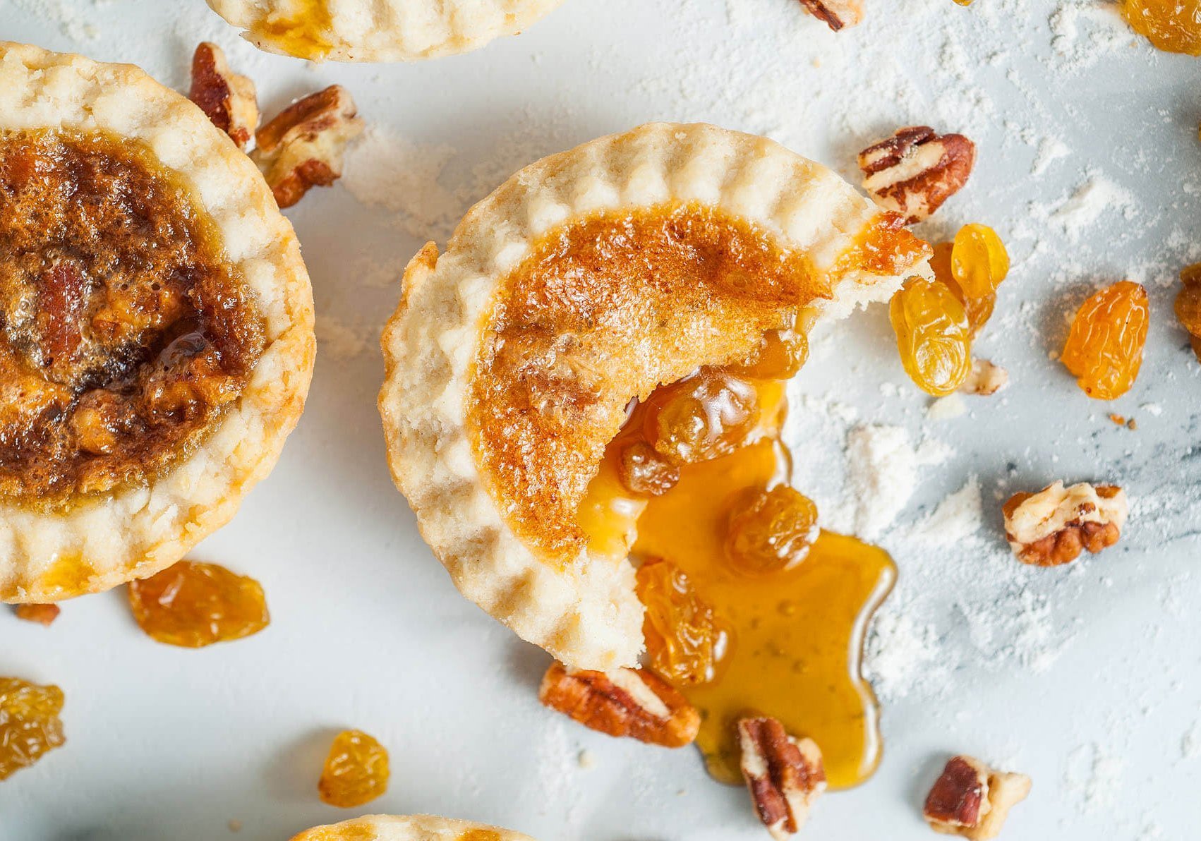 A butter tart with raisins and spilling syrup on a white surface, with raisins, pecans and other tarts around it