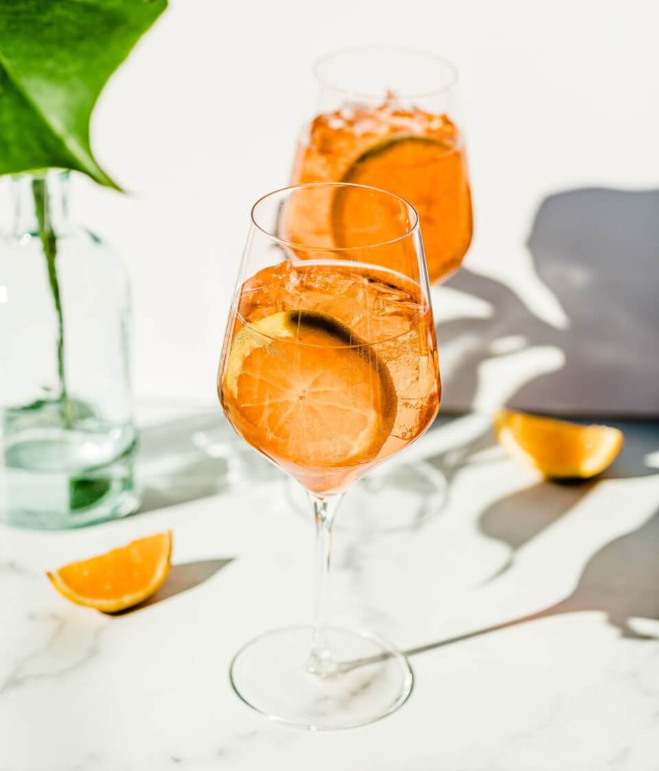 Two orange spritz CBD cocktails with orange wheels on a stark white and sunny backdrop