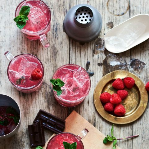 Glasses filled with a red cocktail and raspberries on a grey wooden surface