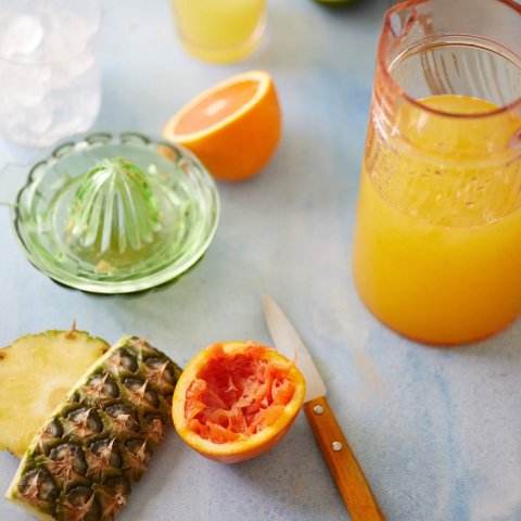 Sliced oranges, pineapples and limes on a white surface with a green glass juicer and pitcher of juice nearby