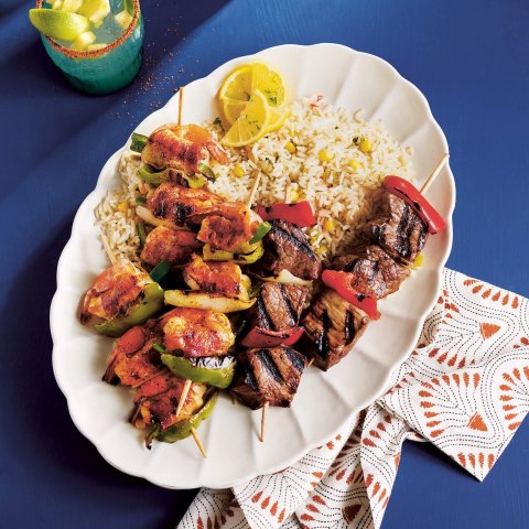 Grilled beef and shrimp skewers with drinks nearby on a bright blue surface