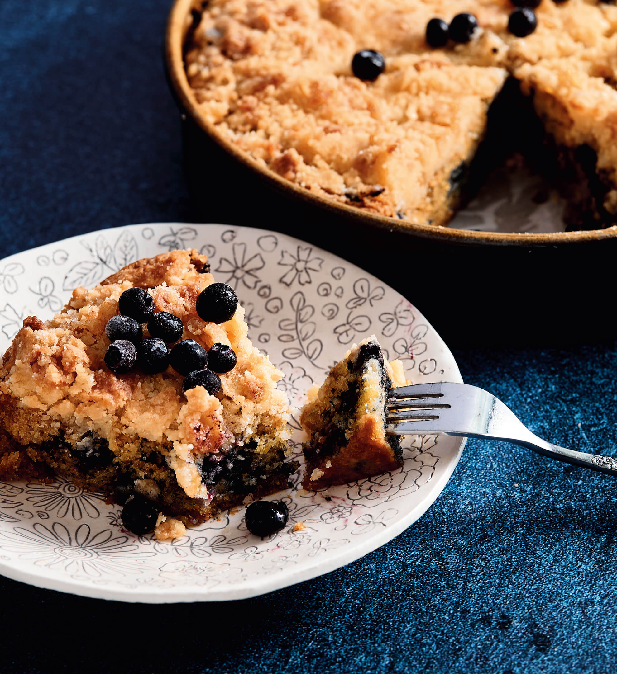 A slice of blueberry crumb cake on a white plate on a blue surface