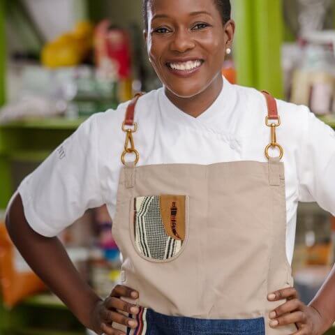 A woman, Selassie Atadika, wears a tan apron and smiles at the camera with her hands on her hips