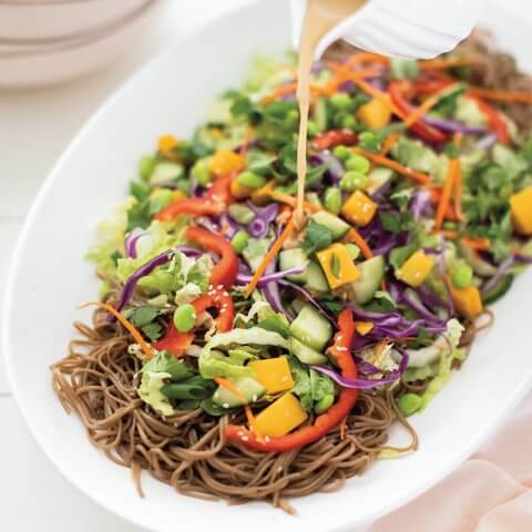 A white dish of soba noodle salad with vegetables and dressing being poured over it