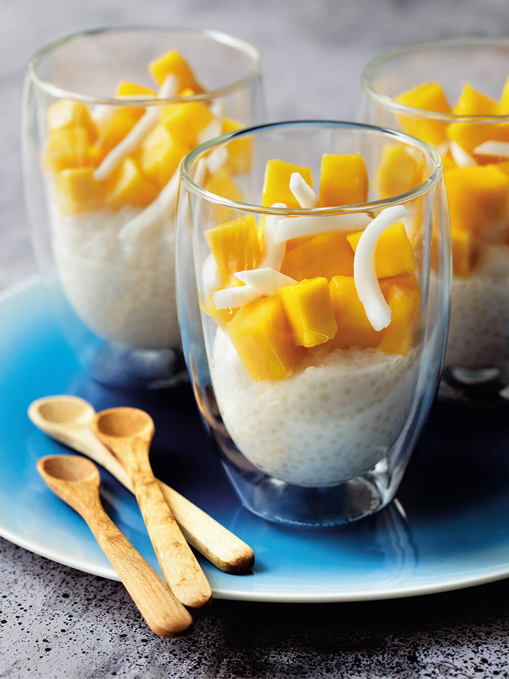 Three glasses with tapioca pudding topped with mango on a blue plate with small wooden spoons