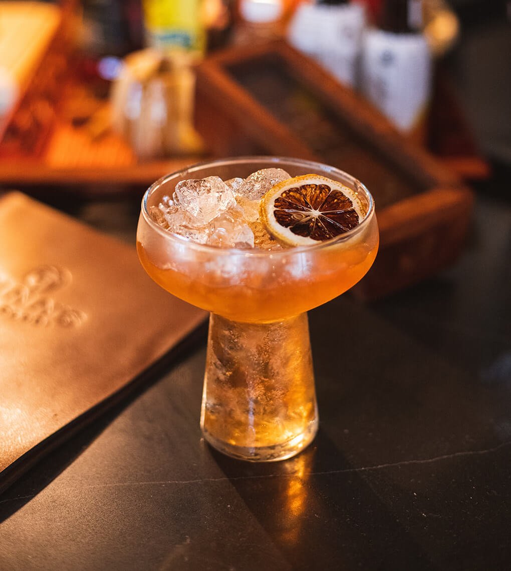 An orange-hued cocktail with a dried citrus garnish on a dark wood table