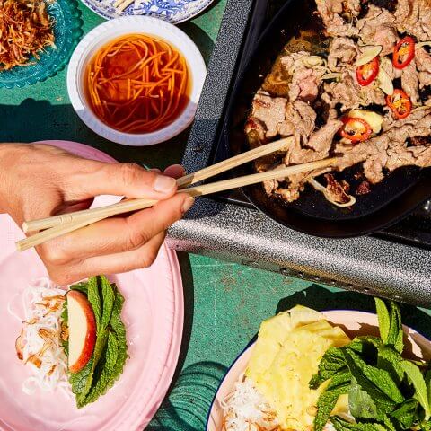A person using chopsticks to take beef off a hot plate at a table, with side dishes and dipping sauce around it