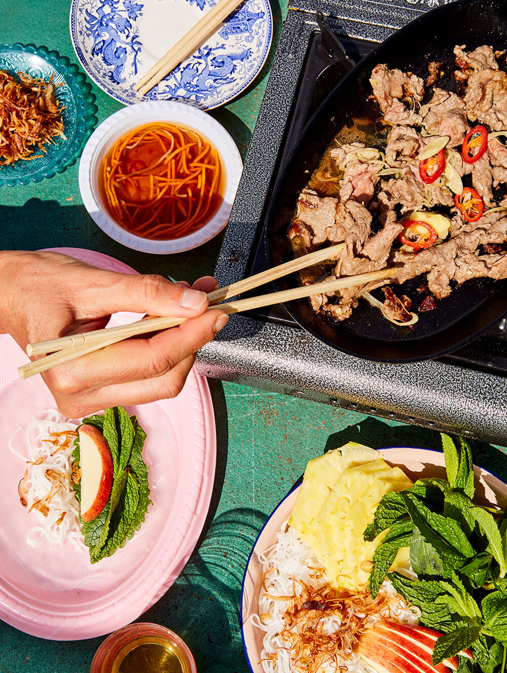 A person using chopsticks to take beef off a hot plate at a table, with side dishes and dipping sauce around it