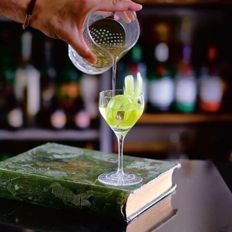 A cocktail being poured into a glass resting on top of a green book