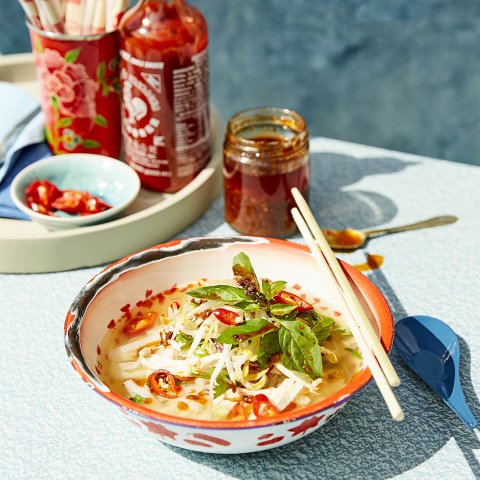 A bowl of chicken pho with sriracha and chopsticks visible in the background